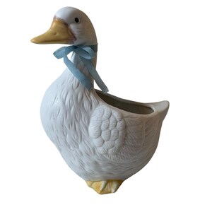 Puede incluir: Una maceta de cerámica blanca con forma de pato, con pico y patas amarillas y detalles de plumas texturizadas en el cuerpo. Una cinta azul claro está atada alrededor del cuello del pato. La parte superior del lomo está abierta y es hueca.