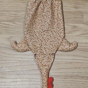Puede incluir: Una bolsa de tela con forma de gallina que presenta un fondo beige con un peque&ntilde;o estampado floral rojo y verde. La bolsa tiene dos patas de tela amarillas en la parte superior, dos alas laterales y una cabeza en la parte inferior con cresta roja, barbilla roja y pico amarillo.
