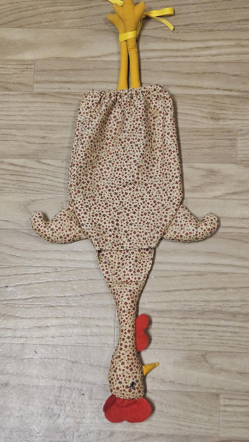 Puede incluir: Una bolsa de tela con forma de gallina que presenta un fondo beige con un peque&ntilde;o estampado floral rojo y verde. La bolsa tiene dos patas de tela amarillas en la parte superior, dos alas laterales y una cabeza en la parte inferior con cresta roja, barbilla roja y pico amarillo.