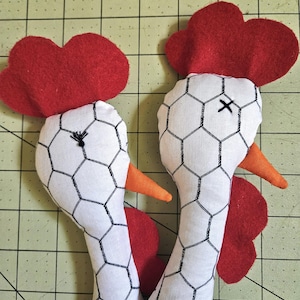 Puede incluir: Dos cabezas de pollo de tela rellena con un patr&oacute;n de malla de alambre negro impreso sobre fondo blanco. Cada una tiene una cresta de fieltro rojo brillante, barbillas de fieltro rojo y un pico de tela naranja. Los ojos est&aacute;n bordados.