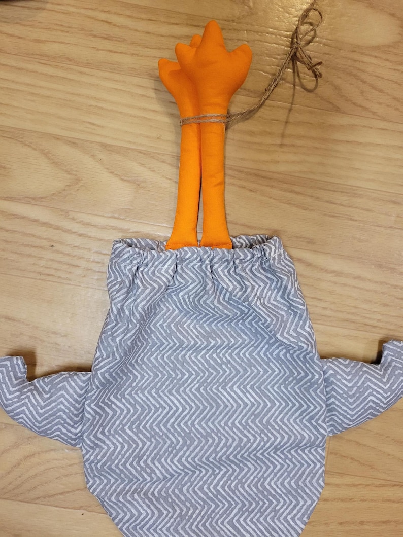 Puede incluir: Una bolsa de tela con un patr&oacute;n de chevr&oacute;n gris y blanco, con dos ap&eacute;ndices naranjas rellenos que sobresalen por la parte superior, asegurada con un cierre de cord&oacute;n y un trozo de cuerda atado alrededor de los elementos naranjas.