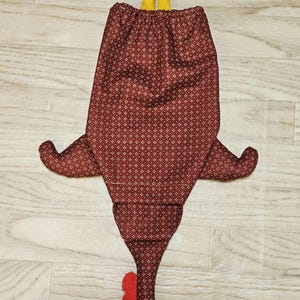 Puede incluir: Una bolsa de tela con forma de pollo en color granate oscuro con un peque&ntilde;o patr&oacute;n geom&eacute;trico blanco. La bolsa presenta patas de tela amarillas en la parte superior, una cresta y barbillas rojas, y un pico amarillo, con una cinta amarilla atada.
