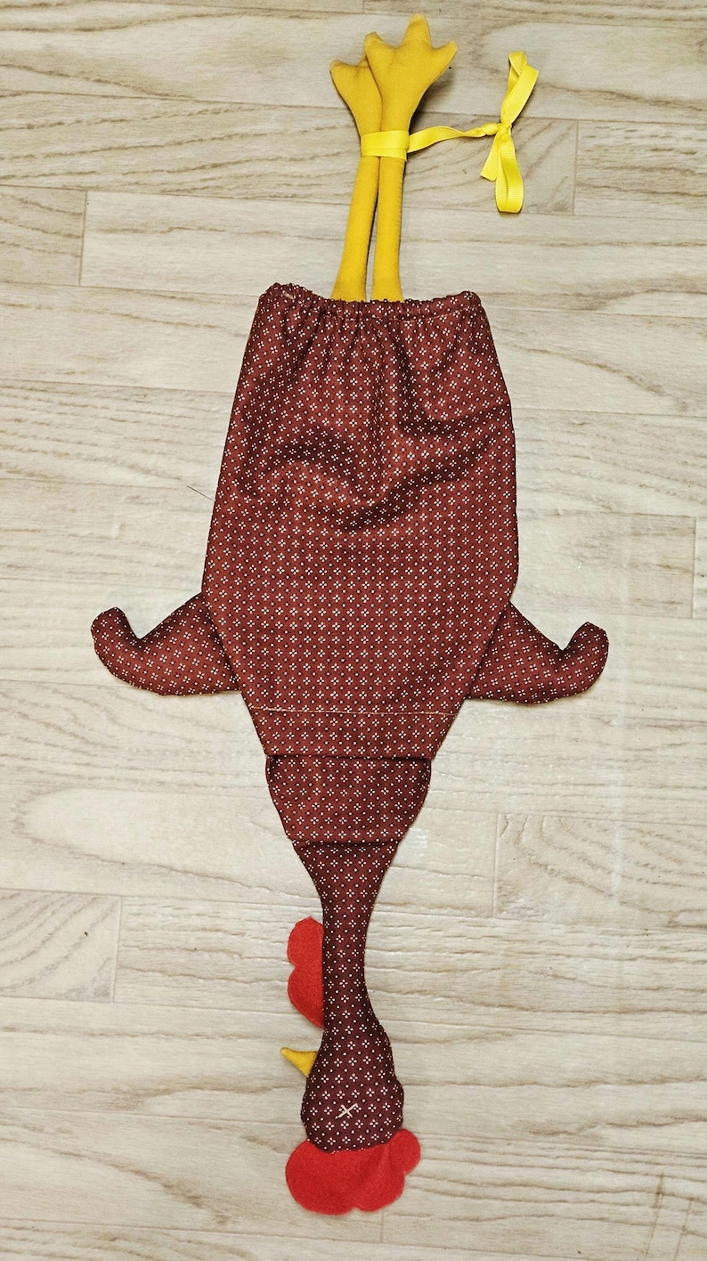Puede incluir: Una bolsa de tela con forma de pollo en color granate oscuro con un peque&ntilde;o patr&oacute;n geom&eacute;trico blanco. La bolsa presenta patas de tela amarillas en la parte superior, una cresta y barbillas rojas, y un pico amarillo, con una cinta amarilla atada.