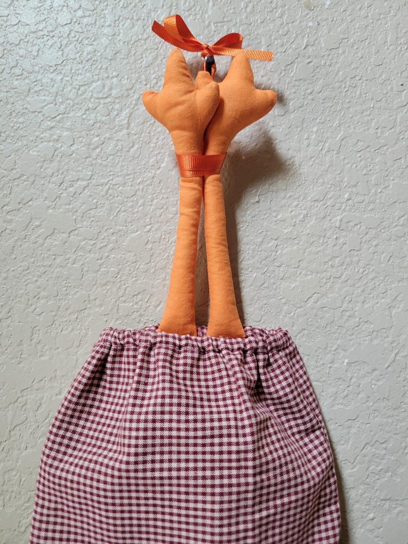 Puede incluir: Una bolsa de tela colgante con un patr&oacute;n de cuadros vichy granate y blanco y una parte superior fruncida. Dos piernas de tela naranja rellenas sobresalen de la abertura, atadas con un lazo de cinta naranja y una banda de cinta a juego.