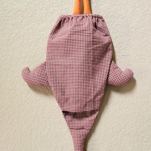 Puede incluir: Una bolsa de almacenamiento de tela con forma de pollo, con un patr&oacute;n de cuadros vichy en color granate y blanco. La bolsa tiene patas de tela naranja en la parte superior con una cinta para colgar y una cabeza de pollo en la parte inferior con cresta roja y pico naranja.