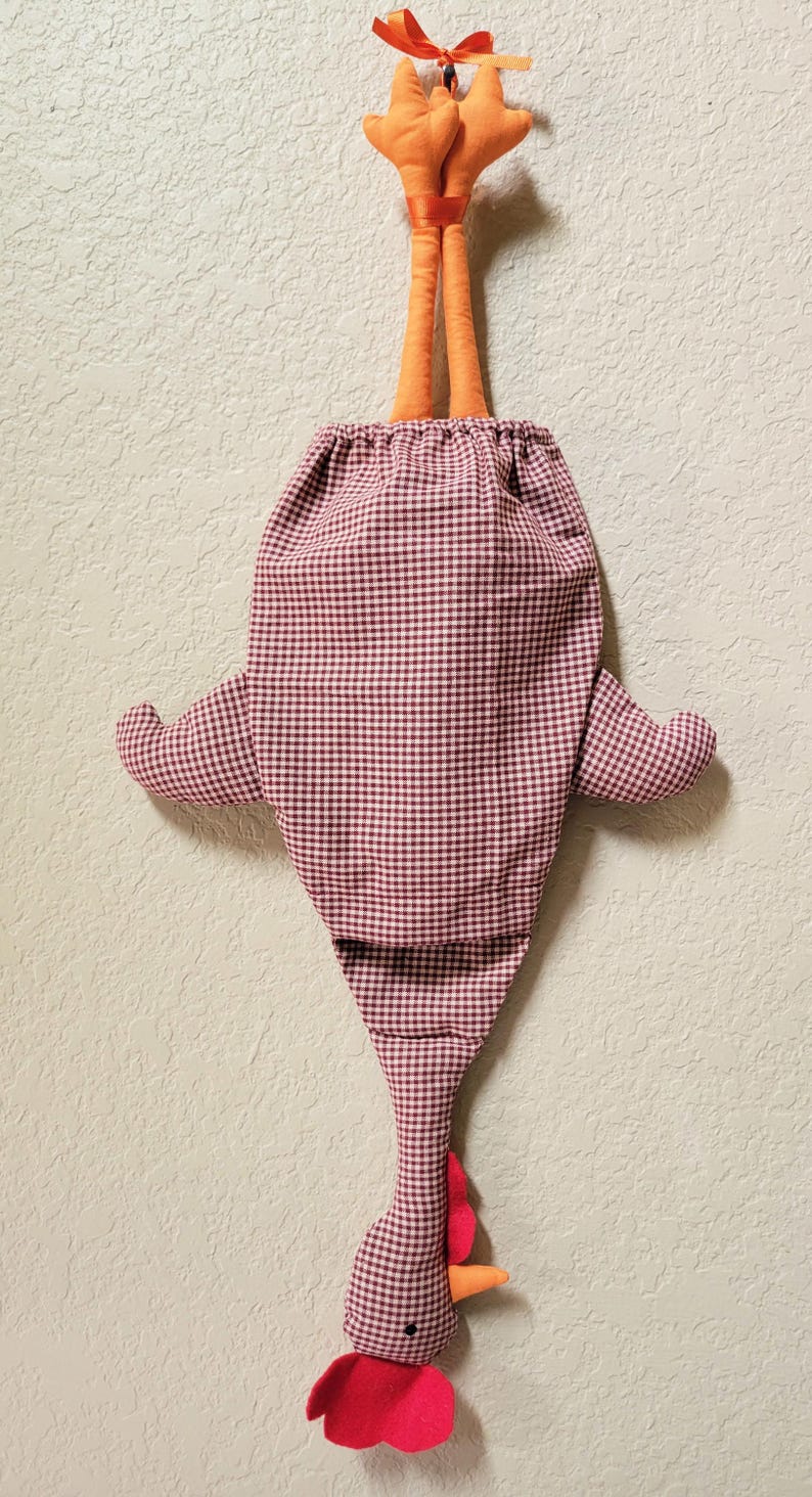 Puede incluir: Una bolsa de almacenamiento de tela con forma de pollo, con un patr&oacute;n de cuadros vichy en color granate y blanco. La bolsa tiene patas de tela naranja en la parte superior con una cinta para colgar y una cabeza de pollo en la parte inferior con cresta roja y pico naranja.