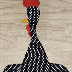 Puede incluir: Un juguete de tela con forma de pollo que presenta un cuerpo negro con lunares blancos, una cresta de fieltro rojo en la parte superior de la cabeza, una barbilla de fieltro rojo bajo el pico y un pico de fieltro amarillo. Tiene cuello largo.