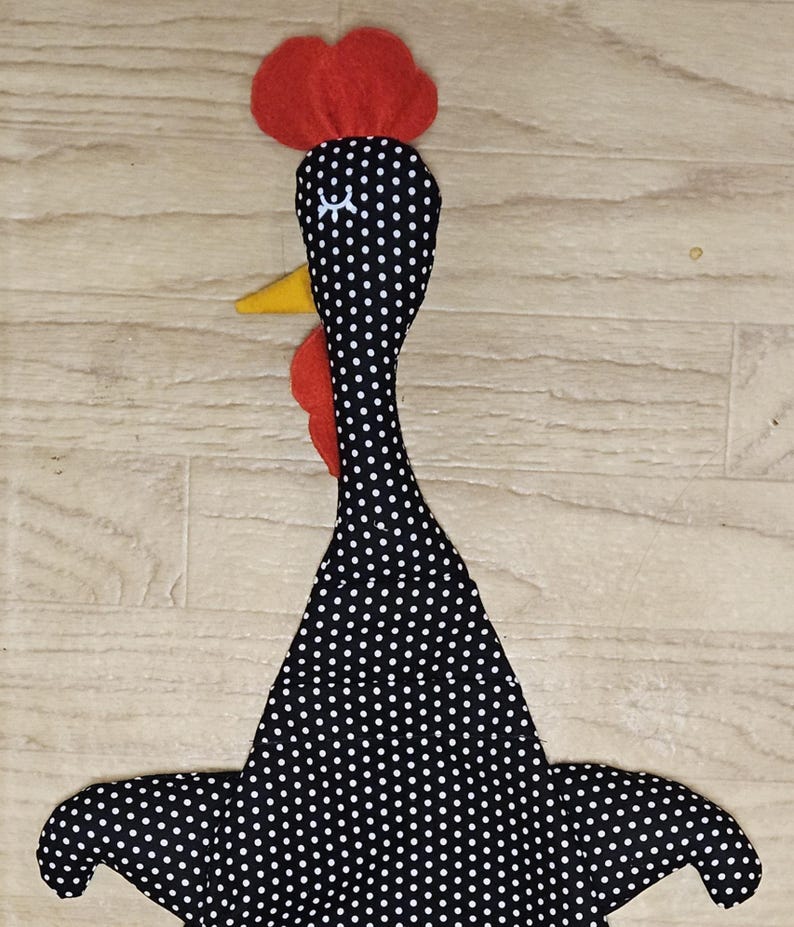 Puede incluir: Un juguete de tela con forma de pollo que presenta un cuerpo negro con lunares blancos, una cresta de fieltro rojo en la parte superior de la cabeza, una barbilla de fieltro rojo bajo el pico y un pico de fieltro amarillo. Tiene cuello largo.