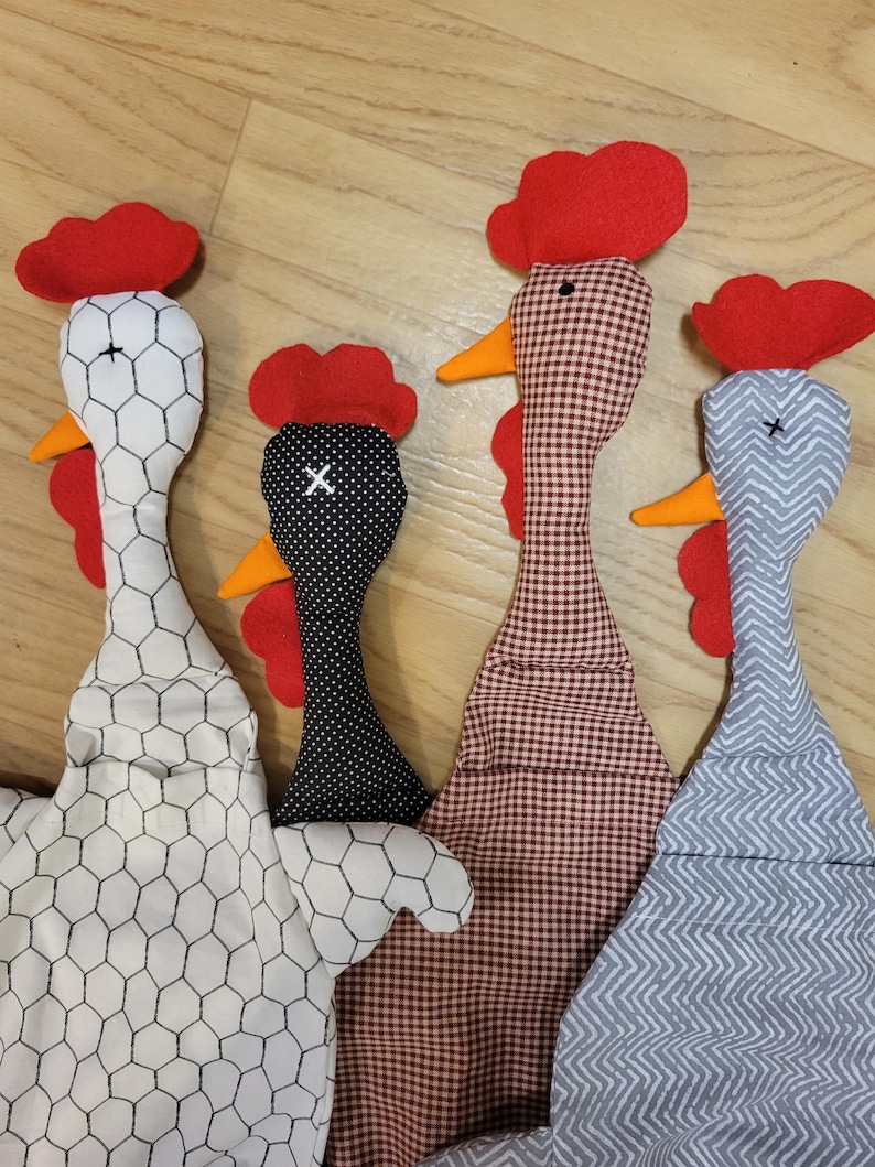 Puede incluir: Cuatro adornos de tela con forma de gallina con distintos patrones: blanco con estampado de malla de gallinero negra, negro con lunares blancos, cuadros vichy marrones y blancos, y gris con patr&oacute;n de chevr&oacute;n blanco. Cada uno tiene cresta y barbillas de fieltro rojo.