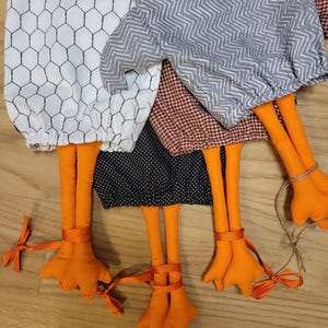 Puede incluir: Cuatro adornos de tela con forma de pollo con varios patrones, incluyendo blanco con malla negra, chevron gris, cuadros vichy marrones y negro con lunares blancos. Cada adorno tiene patas largas de color naranja con lazos atados.