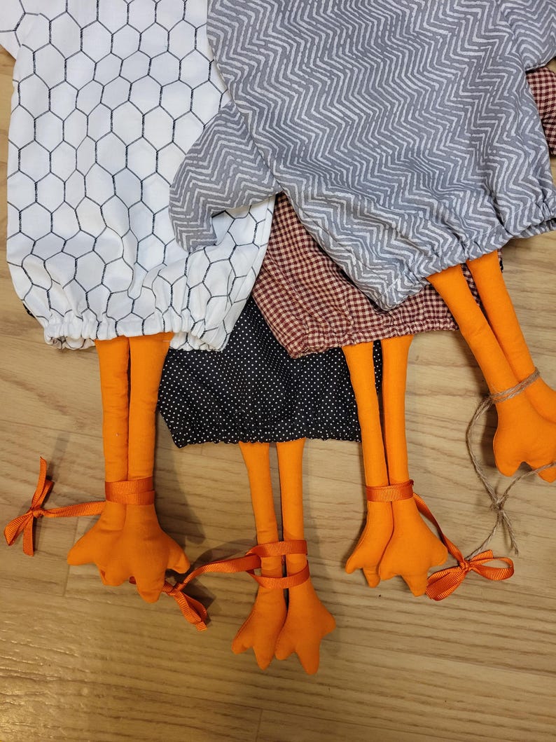 Puede incluir: Cuatro adornos de tela con forma de pollo con varios patrones, incluyendo blanco con malla negra, chevron gris, cuadros vichy marrones y negro con lunares blancos. Cada adorno tiene patas largas de color naranja con lazos atados.