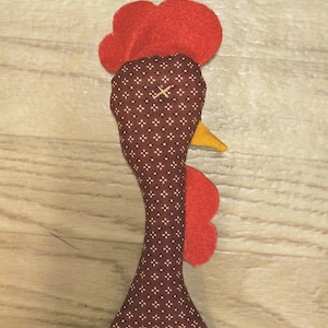 Puede incluir: Un adorno de tela con forma de cabeza de gallo, confeccionado en tela marr&oacute;n con un peque&ntilde;o patr&oacute;n geom&eacute;trico blanco, cresta y barbillas de fieltro rojo brillante y un peque&ntilde;o pico de fieltro amarillo.