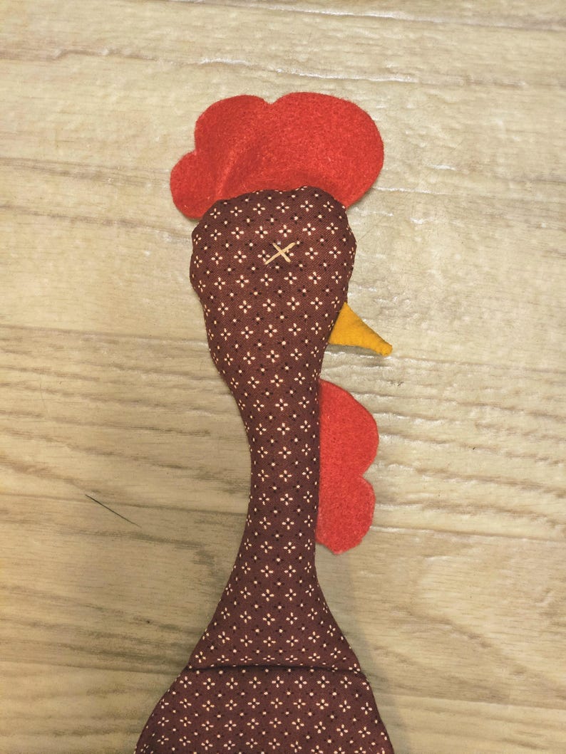 Puede incluir: Un adorno de tela con forma de cabeza de gallo, confeccionado en tela marr&oacute;n con un peque&ntilde;o patr&oacute;n geom&eacute;trico blanco, cresta y barbillas de fieltro rojo brillante y un peque&ntilde;o pico de fieltro amarillo.