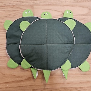 May include: Three turtle-shaped fabric items featuring dark green circular shells with a cross-stitched pattern. Each piece has a light green head with two white eyes, four light green flippers, and a pointed tail. The underside is textured.