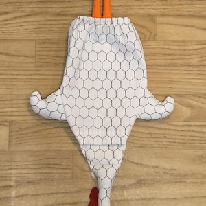 Puede incluir: Una bolsa de tela blanca con forma de pollo y un patr&oacute;n de malla hexagonal negra. La bolsa presenta patas de tela naranja en la parte superior, una cresta y barbillas rojas, y un pico naranja en la parte inferior. Incluye un cierre de cord&oacute;n.