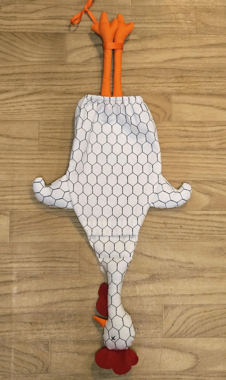 Puede incluir: Una bolsa de tela blanca con forma de pollo y un patr&oacute;n de malla hexagonal negra. La bolsa presenta patas de tela naranja en la parte superior, una cresta y barbillas rojas, y un pico naranja en la parte inferior. Incluye un cierre de cord&oacute;n.