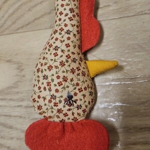 Puede incluir: Un juguete de tela relleno con forma de pollo, con una base de color crema y un peque&ntilde;o estampado floral rojo y azul. Tiene un pico de fieltro amarillo brillante, una cresta roja en la parte superior, barbillas rojas y un ojo cosido en negro.