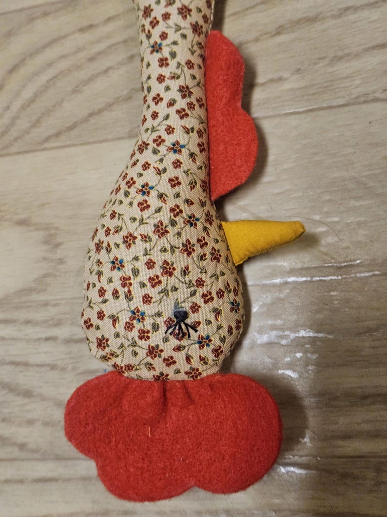 Puede incluir: Un juguete de tela relleno con forma de pollo, con una base de color crema y un peque&ntilde;o estampado floral rojo y azul. Tiene un pico de fieltro amarillo brillante, una cresta roja en la parte superior, barbillas rojas y un ojo cosido en negro.