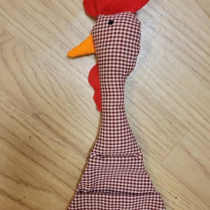 Puede incluir: Un juguete de tela con forma de gallo que presenta un patr&oacute;n de cuadros vichy en marr&oacute;n y blanco en la cabeza y el cuello. El gallo tiene una cresta y barbillas de fieltro rojo brillante, un pico naranja y un ojo circular negro.