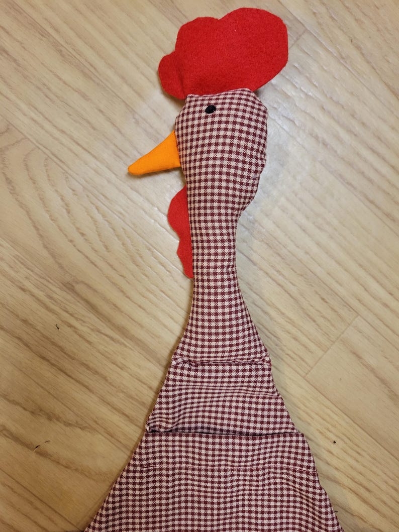Puede incluir: Un juguete de tela con forma de gallo que presenta un patr&oacute;n de cuadros vichy en marr&oacute;n y blanco en la cabeza y el cuello. El gallo tiene una cresta y barbillas de fieltro rojo brillante, un pico naranja y un ojo circular negro.
