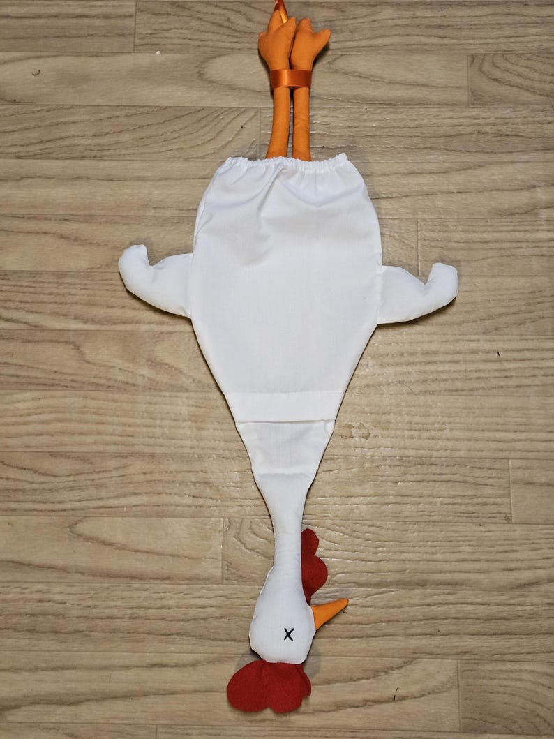 Puede incluir: Una bolsa de tela blanca con forma de pollo y una abertura el&aacute;stica en la parte superior. La bolsa presenta patas naranjas que sobresalen por arriba, una cabeza blanca con cresta y barbillas rojas, un pico naranja y una X negra bordada.