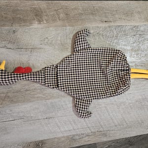 Puede incluir: Un accesorio de cocina de tela con forma de pollo y estampado de cuadros vichy en blanco y negro. La figura tiene una cresta y barbillas de fieltro rojo brillante, un pico amarillo y dos patas de fieltro amarillo en la parte trasera.