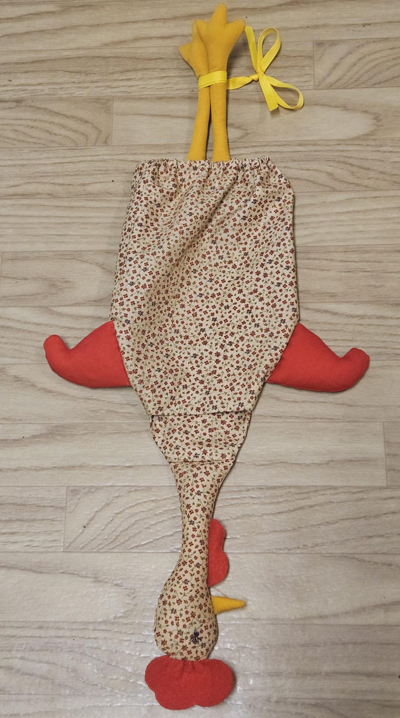 Puede incluir: Una bolsa de tela con forma de gallina con un cuerpo de color crema que presenta un peque&ntilde;o estampado floral rojo y azul. Tiene dos patas de tela amarilla en la parte superior, alas rojas en los lados, y una cresta roja y un pico amarillo.
