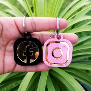 May include: Two square keychains with rounded corners attached to silver metal rings. One keychain is black with a gold-toned circular center featuring a black Facebook logo. The other is light pink with a gold-toned square frame and center.