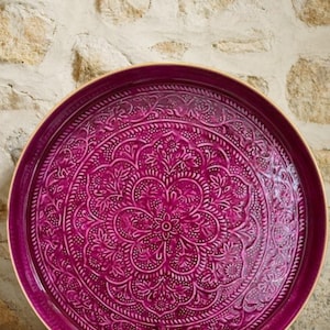 Peut inclure: Un plateau de service rond magenta avec un motif floral et géométrique complexe. Le plateau a un bord doré et un motif en relief. Le plateau est probablement en céramique ou dans un matériau similaire.