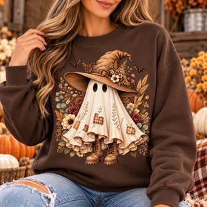 May include: A dark brown crewneck sweatshirt featuring a central graphic of a ghost wearing a large, wide-brimmed witch hat adorned with flowers. The ghost is wearing brown lace-up boots and is surrounded by autumn-themed foliage and small bats.