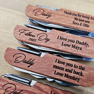 May include: Close-up of several personalized wooden multi-tools. Each tool has a wooden handle with engraved text, including "Daddy," "Fathers Day 2025," and loving messages from children. The tools are made of stainless steel.
