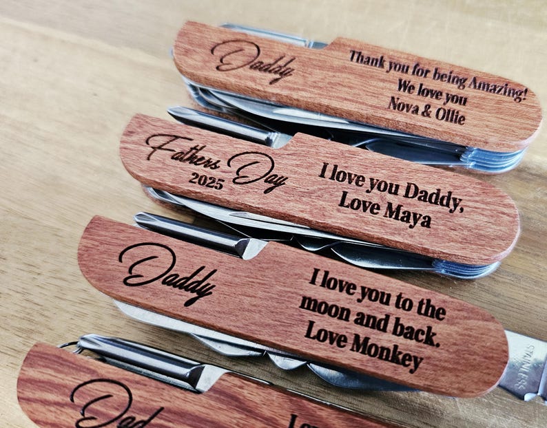 May include: Close-up of several personalized wooden multi-tools. Each tool has a wooden handle with engraved text, including "Daddy," "Fathers Day 2025," and loving messages from children. The tools are made of stainless steel.