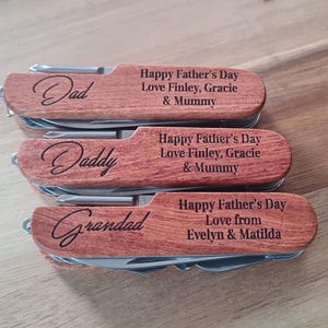 Può includere: Tre coltelli tascabili in legno personalizzati con utensili in acciaio inossidabile. Ogni coltello è inciso con un nome diverso: "Dad", "Daddy" e "Grandad". I coltelli presentano anche il testo "Happy Father's Day" e un messaggio personalizzato.