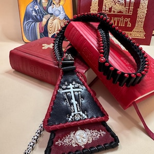 Può includere: Una corda di preghiera in pelle nera e rossa con un ciondolo a forma di croce, appoggiata su libri rossi e un'icona. La corda di preghiera presenta perline rosse e un design intrecciato. Il ciondolo ha una croce bianca.