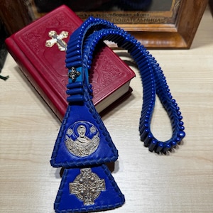 May include: A blue leather prayer rope with silver religious icons and a cross, resting on a red book with a gold cross. The prayer rope is made of blue beads and leather. A framed religious icon is in the background.