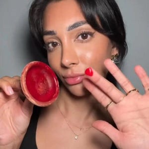 Può includere: Primo piano di un prodotto per labbra rosso tenuto in mano da una persona. Il prodotto è in un contenitore rotondo di colore chiaro. La persona ha lo smalto rosso su un'unghia e indossa anelli d'oro e una collana.