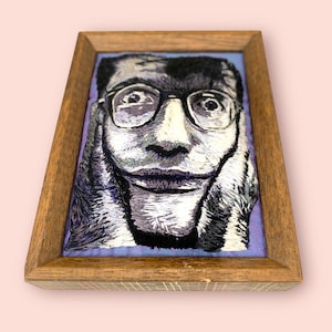 May include: Embroidered portrait of a person wearing glasses, framed in a wooden frame. The artwork is primarily black and white, with details in shades of gray and purple. The person's face is the central focus of the artwork.