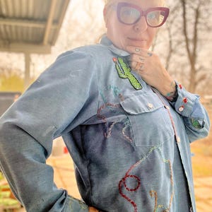 May include: Light blue denim shirt with colorful embroidered details. The shirt features a green cactus patch and intricate stitching in red, yellow, and brown. The cuffs and collar are also embellished with embroidery, creating a unique, handcrafted look.