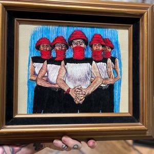 May include: Framed artwork featuring an embroidered image of five figures. Each figure wears a red hat and scarf, black and white top, and black pants. The background is blue. The frame is gold-toned with a black inner border.