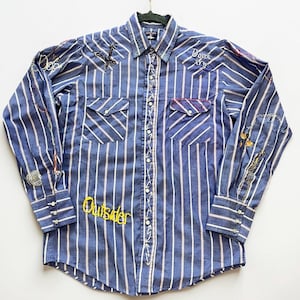 May include: Blue and white striped button-down shirt with embroidered details. Features a cross and the word "Outsider" in yellow. The shirt has two chest pockets and is hanging on a black hanger.