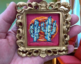 Peut inclure: Un petit cadre doré orné de motifs élaborés. Il encadre une œuvre d'art brodée miniature représentant des cactus bleus sur un fond orange et rose. Le cadre est tenu dans une main, soulignant la taille et les détails de l'œuvre.