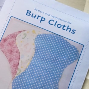 May include: A pattern and instructions for burp cloths. The image shows three burp cloths in different colors and patterns: pink, white with small designs, and blue with white polka dots. The text is in blue.