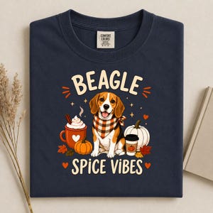 May include: A navy blue t-shirt featuring a graphic of a beagle wearing a plaid bandana, surrounded by a mug of whipped cream, a pumpkin, and a coffee cup. The text reads BEAGLE SPICE VIBES, accented with small hearts, stars, and autumn leaves.