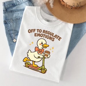 May include: White t-shirt with the text "OFF TO REGULATE EMOTIONS" in brown. The shirt features a cartoon duck riding a scooter and holding a coffee cup. The duck has a bow tie and is surrounded by yellow stars. The shirt is paired with blue jeans and a brown hat.