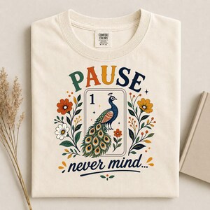 Könnte beinhalten: Ein cremefarbenes T-Shirt mit einem Grafikdruck, der den Text PAUSE und never mind zeigt. Das Design enthält eine zentrale Spielkartenillustration eines Pfaus mit der Zahl 1, umgeben von farbenfrohen Blumen- und Blattmotiven.