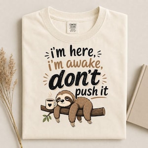 Pode incluir: Uma t-shirt de cor creme com um gráfico de uma preguiça castanha pendurada num ramo com uma caneca de café a fumegar. O texto diz "i'm here, i'm awake, don't push it" em letras pretas e castanhas. A etiqueta Comfort Colors é visível.