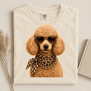May include: A cream-coloured t-shirt featuring a graphic of a curly-haired poodle wearing leopard-print sunglasses, a matching leopard-print neck scarf, and a thick gold-tone chain necklace. The inner neck label displays the text Comfort Colors.