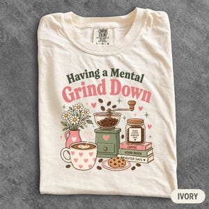 May include: An ivory t-shirt featuring a graphic with the text Having a Mental Grind Down. The design includes a coffee grinder, a mug with hearts, a jar of coffee beans, a stack of books labelled Coffee, Good Mood, Brighter Days, and flowers.