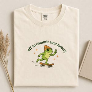 Könnte beinhalten: Ein cremefarbenes T-Shirt mit einer Grafik eines grünen Frosches, der eine orangefarbene Mütze trägt, Skateboard fährt und ein Friedenszeichen macht. Der Text "off to commit aunt foolery" steht in Dunkelgrün darüber, ergänzt durch orange Sterne.