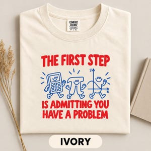 May include: An ivory t-shirt featuring red text that reads THE FIRST STEP IS ADMITTING YOU HAVE A PROBLEM. Below the text are blue line-art illustrations of a smiling calculator, the pi symbol with arms and legs, and a graph with a smiling face.